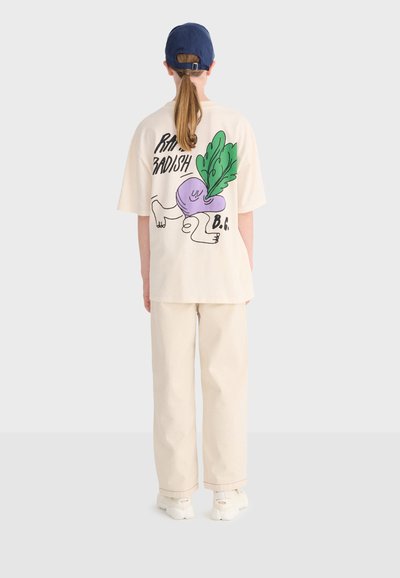 Person iført cremefarvede bukser og en oversize creme t-shirt med en farverig cartoon af en radise og tekst på ryggen, kombineret med hvide sko og en blå kasket.