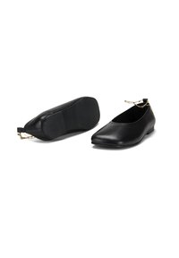 Een paar zwarte leren balletschoenen met puntige neuzen en gouden enkelkettinkjes, één schoen rechtop en één op zijn zijkant.