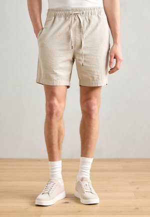 Homme portant un short beige texturé, des chaussettes blanches et des baskets beige clair à lacets, debout sur un sol en bois.