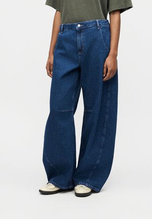 Personne portant un jean bleu foncé à jambes larges avec des coutures visibles, associé à des chaussures beiges et une chemise verte à manches courtes, debout.