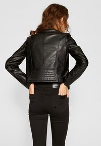 Kobieta z brązowymi włosami zebranymi w kok, ubrana w czarną skórzaną kurtkę i czarne spodnie, stoi z rękami w tylnej kieszeni, zwrócona plecami.