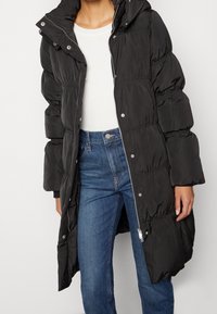 Black puffer coat with a hood, featuring a zip closure and snap buttons. The coat has a quilted texture and a long, relaxed fit.