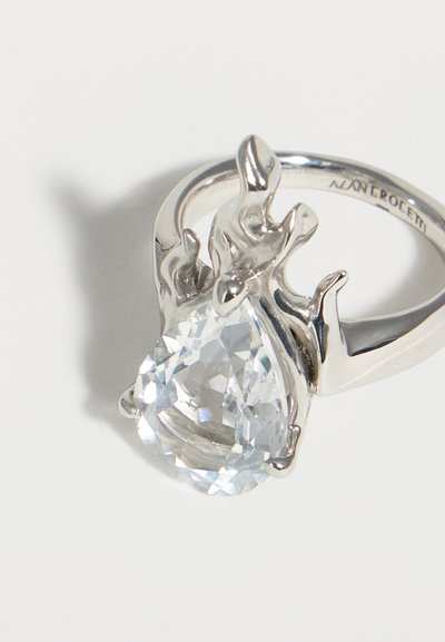 Ασημένιο δαχτυλίδι με ένα αχλαδόσχημο, διαυγή πολύτιμο λίθο που κρατιέται από ένα αφηρημένο μεταλλικό σχέδιο σε σχήμα φωτιάς σε μία λεία μπάντα.