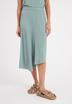 Vrouw draagt een lichtgroene asymmetrische midirok gecombineerd met beige touwsandalen tegen een witte achtergrond.