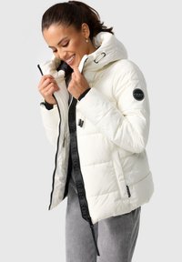 White puffy jacket with a high collar, front zipper, and logo patch on the sleeve; features black zipper trim and adjustable hood.