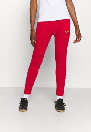 Person, die rote Nike Trainingshose, weiße Socken, schwarze Adidas Schuhe und ein weißes Oberteil trägt, steht auf einem hellgrauen Boden vor einer weißen Wand.