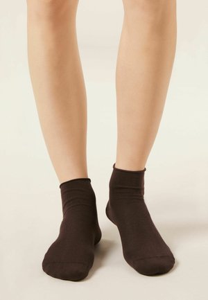 Blote benen met effen donkerbruine enkelsokken, staand op een lichtgekleurde vloer tegen een neutrale achtergrond.