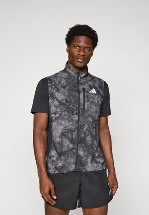 Athletic man wearing black patterned zip-up vest over black short-sleeve shirt and black shorts with sports brand logos, standing against white background.