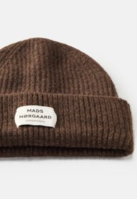 Brun strikket lue med ribbet tekstur, som har et hvitt merke med teksten "MADS NØRGAARD COPENHAGEN" på bretten. Mykt materiale.