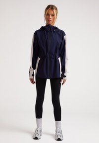 Chaqueta con capucha en azul marino y blanca, con cintura ajustada, cierre de cremallera y bolsillos laterales, combinada con leggins negros y zapatillas blancas.