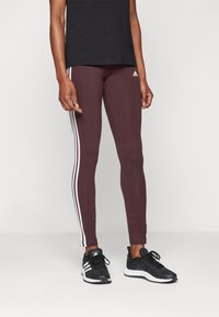Person i vinröda Adidas leggings med vita ränder och svarta Adidas sneakers, som står mot en enkel ljus bakgrund.