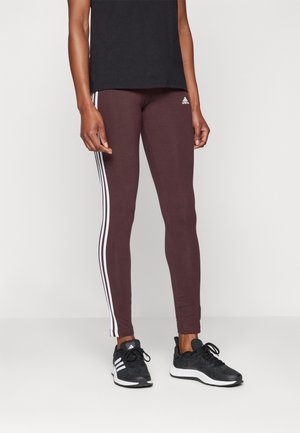 Persona vistiendo leggings burdeos de Adidas con rayas blancas y zapatillas negras de Adidas, de pie contra un fondo claro y liso.
