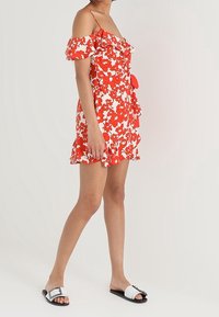 Röd blommig klänning med avskuren axel, volangärmar och volang i nederkant, med en figurnära byst och dekoration i form av en rosett. Bärs med svartvita sandaler.