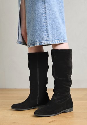 Bottes noires en suede au-dessus du genou avec un design froncé, semelle plate et fermeture éclair sur le côté; portées avec une jupe en denim bleu clair sur un sol en bois.