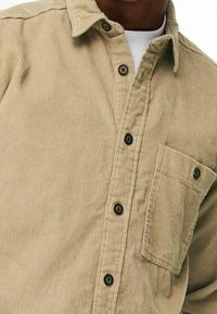 Beige Cord-Shirt mit Knopfverschluss und Brusttasche, über einem weißen Rundhalsausschnitt-Shirt getragen.
