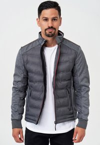 Veste isolante grise avec un design matelassé, des manches texturées, des accents rouges, un col haut et deux poches zippées. Portée par-dessus un t-shirt blanc.