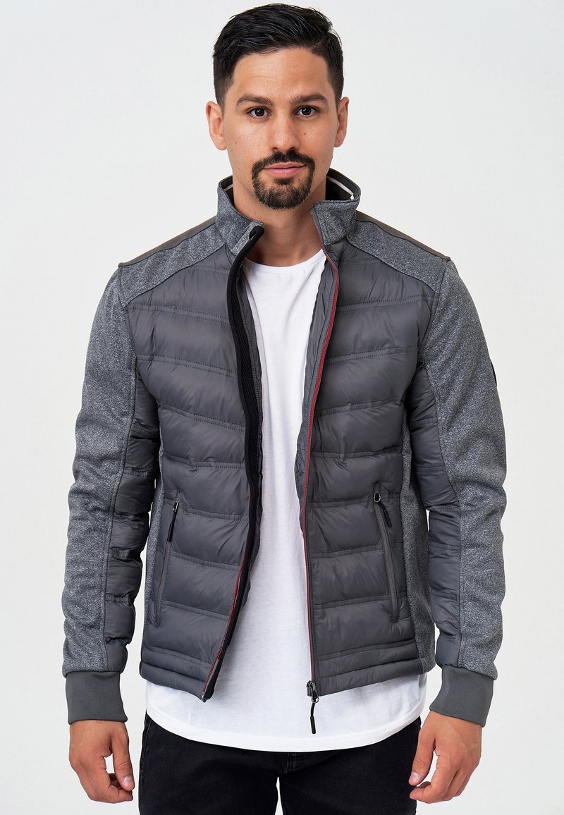 Veste isolante grise avec un design matelassé, des manches texturées, des accents rouges, un col haut et deux poches zippées. Portée par-dessus un t-shirt blanc.