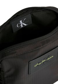 Bolsa de tela negra con cierre de cremallera, que presenta un interior suave, etiqueta con el logo de CK y una etiqueta rectangular con "Calvin Klein Jeans" en verde lima.