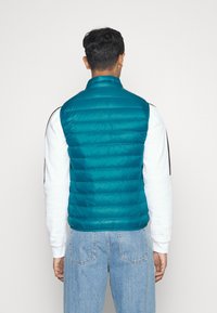 Turkusowa kamizelka puchowa z wysokim kołnierzem i pikowanym wzorem, zestawiona z białą koszulą z długim rękawem i jasnoniebieskimi dżinsami. Prosty design, bez widocznych logo.