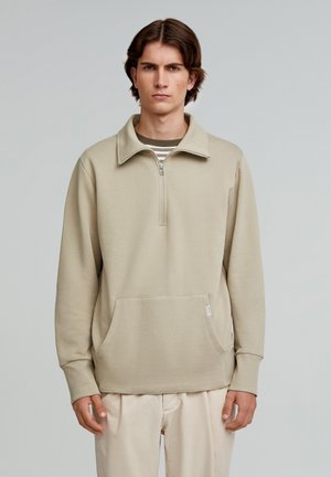 Beige zip-up sweatshirt with a high collar, a front pocket, textured fabric, and long sleeves, worn over a striped shirt.