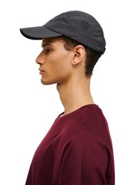 Junger Mann mit dunkelgrauer Kappe und burgunderrotem Hemd, im Profil vor einem schlichten weißen Hintergrund.