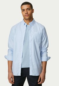 Lichtblauw overhemd met lange mouwen, met borstzak, gedragen over een lichtblauwe t-shirt, gecombineerd met donkere broek. Gladde stofstructuur.
