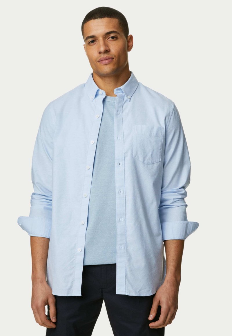 Lichtblauw overhemd met lange mouwen, met borstzak, gedragen over een lichtblauwe t-shirt, gecombineerd met donkere broek. Gladde stofstructuur.