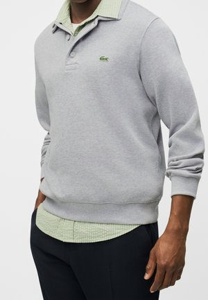 Homme portant un sweat-shirt polo manches longues gris clair sur une chemise texturée vert pâle avec un pantalon noir, mains dans les poches.