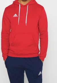 Red hoodie with a front kangaroo pocket, white drawstrings, and an embroidered white Adidas logo on the left chest. Cotton blend fabric.