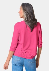 Magentafarbenes Oberteil mit 3/4-Ärmeln, lockerer Passform und abgerundetem Saum. Getragen mit blauen Jeans, die über eine helle Stickerei auf den Gesäßtaschen verfügen.