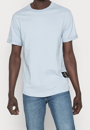 Person trägt ein hellblaues kurzärmeliges T-Shirt mit einem kleinen schwarzen Logopatch auf der unteren linken Seite, kombiniert mit blauen Jeans.
