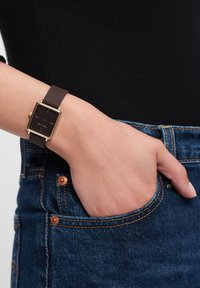 Quadratische goldfarbene Uhr mit schwarzem Zifferblatt, braunem Silikonarmband und minimalistischem Design. Am Handgelenk neben Denim-Stoff getragen.