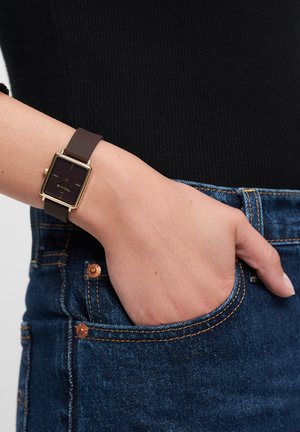 Square gold-tone watch with a black dial, brown silicone strap, and minimalist design. Worn on a wrist beside denim fabric.
