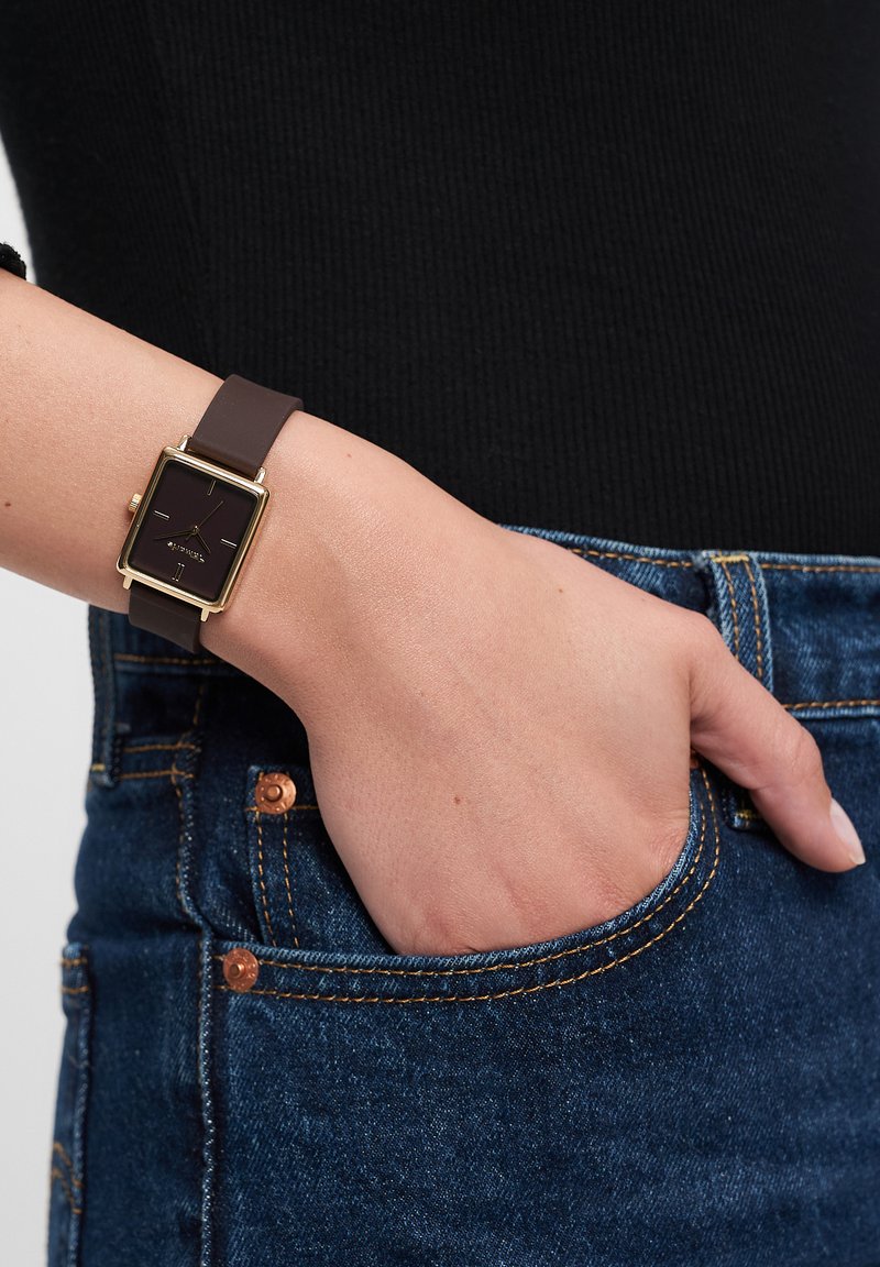 Quadratische goldfarbene Uhr mit schwarzem Zifferblatt, braunem Silikonarmband und minimalistischem Design. Am Handgelenk neben Denim-Stoff getragen.