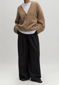 Beżowy sweter z dzianiny z dekoltem w serek, pięcioma guzikami, prążkowanymi mankietami, noszony na białej koszuli, w połączeniu z szerokimi czarnymi spodniami i czarnymi butami.