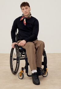 Mand i sort sweater, brune bukser og mønstret tørklæde, siddende i en sort kørestol på en beige gulv foran en ensfarvet væg.