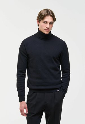 Junger Mann mit hellbraunen Haaren, der einen schwarzen Rollkragenpullover und schwarze Hosen vor einem schlichten hellen Hintergrund trägt.