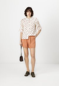 White blouse with red floral print, short flared sleeves; paired with orange shorts. Model holding a small black handbag and wearing green shoes.