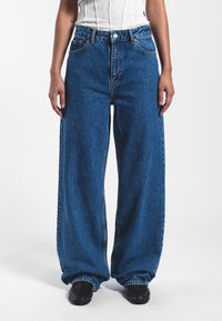 Jeans de mezclilla azul de pierna ancha con una cintura alta, que cuentan con un cierre de botón, bolsillos frontales y una textura ligeramente descolorida.