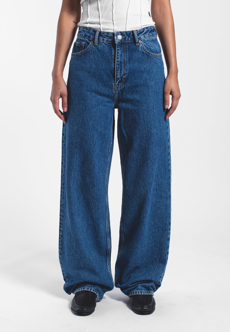 Jeans de mezclilla azul de pierna ancha con una cintura alta, que cuentan con un cierre de botón, bolsillos frontales y una textura ligeramente descolorida.