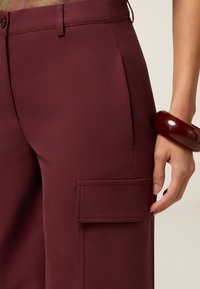 Burgundowe spodnie cargo wykonane z gładkiego materiału, z boczną kieszenią i eleganckim pasem, dopełnione błyszczącym, ciemnym pierścieniem noszonym na nadgarstku.