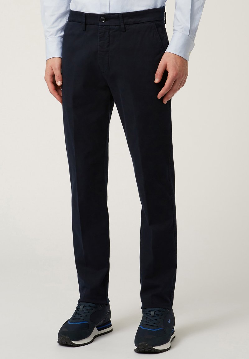 Temno modri chinos hlače iz bombaža. Slim fit z stranskimi in zadnjimi žepi, vsebujejo zapiranje na gumb in zanke za pas. Uporabljeni s temnimi športnimi čevlji.