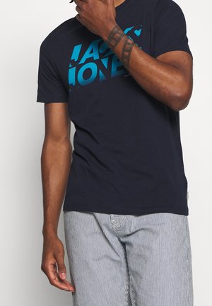 Homme portant un t-shirt noir avec le texte bleu "JACK & JONES" et un pantalon gris clair à rayures, montrant un avant-bras tatoué, sur un fond blanc.