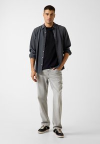 Graues Hemd über einem schwarzen T-Shirt, kombiniert mit hellgrauen Jeans und schwarzen Sneakers mit cremefarbenen Akzenten. Einfaches, modernes Design.