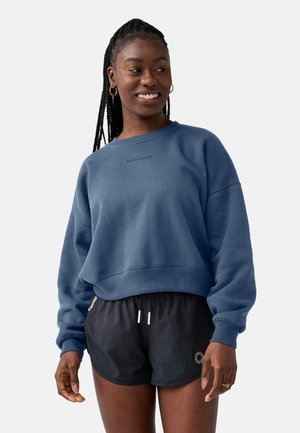 Lachende vrouw met lang gevlochten haar, gekleed in een blauw sweatshirt en zwarte sportshort, staand tegen een effen achtergrond.