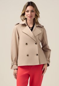 Beige dubbelux jacket met gouden knopen en schouderepauletten. Heeft een gestructureerde vorm en manchetten met bandjes. Gekoeld met rode broeken.