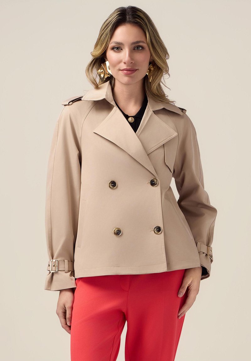 Beige dubbelux jacket met gouden knopen en schouderepauletten. Heeft een gestructureerde vorm en manchetten met bandjes. Gekoeld met rode broeken.