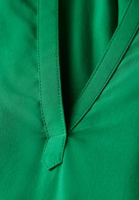 Tessuto verde con scollatura a V e dettagli di cucitura visibili. Il materiale appare liscio con una leggera lucentezza. Nessun motivo o accento.