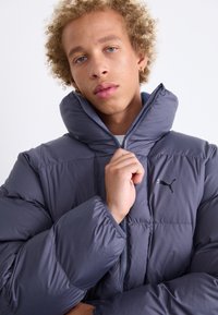 Giacca a vento grigia con collo alto, tessuto lucido, chiusura con zip, texture trapuntata, con un piccolo logo nero Puma sul lato sinistro.