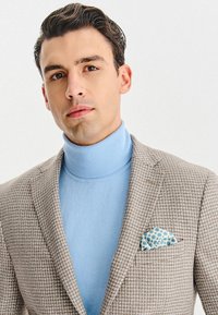 Brązowa kratkowana marynarka na jasnoniebieskim golfie, z wzorzystą poszetką w niebieskim i białym kolorze. Gładki, dopasowany kroj.